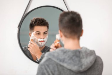 Yakışıklı genç adam banyoda aynanın yanında tıraş oluyor.