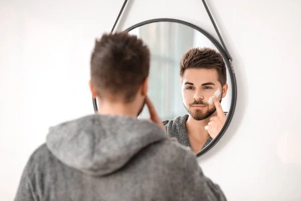 Όμορφος νεαρός που βάζει κρέμα στο πρόσωπό του κοντά στον καθρέφτη στο μπάνιο. — Φωτογραφία Αρχείου