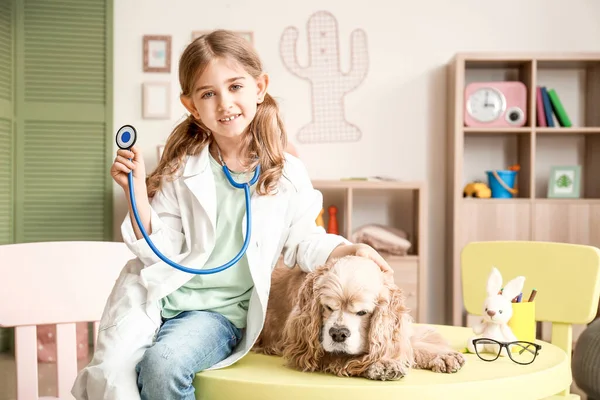 Cute little girl dressed as doctor playing with dog at home — Stock Photo, Image