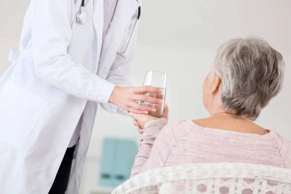 Médico mujer que da a la mujer mayor que sufre de síndrome de Parkinson vaso de agua en la clínica —  Fotos de Stock