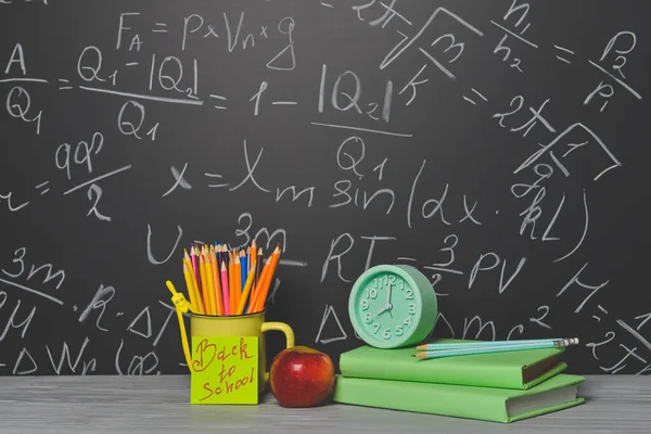 Schulbedarf, Apfel und Uhr auf dem Tisch neben der Tafel — Stockfoto