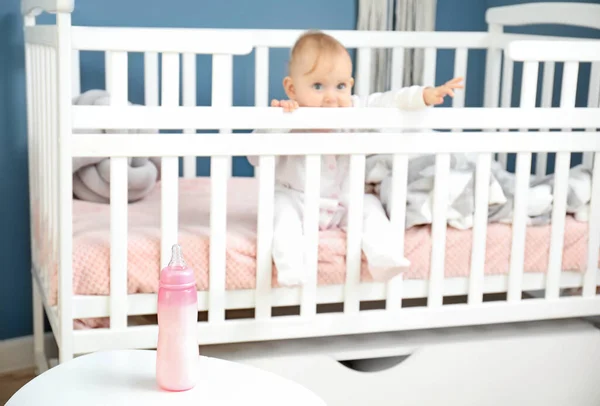 Cute baby sitting in crib near bottle of milk on table — Stock Photo, Image