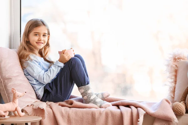 Cute little girl drinking hot chocolate on window sill at home — Stock Photo, Image