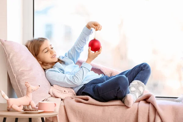 Cute little girl with Christmas ball on window sill at home — Stock Photo, Image