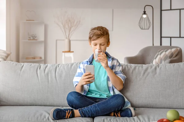 Cute little boy with mobile phone drinking water at home — Stock Photo, Image