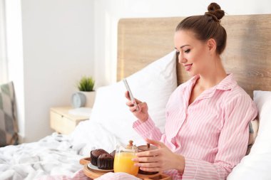 Morning of beautiful young woman with mobile phone having breakfast in bed