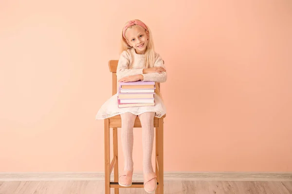 Kleines Mädchen mit Büchern sitzt auf Stuhl neben Farbwand — Stockfoto