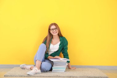 Young woman with books sitting near color wall