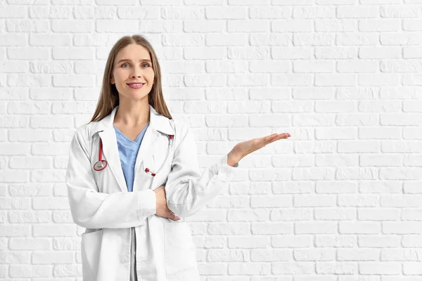 Retrato de médico feminino mostrando algo no fundo de tijolo branco — Fotografia de Stock