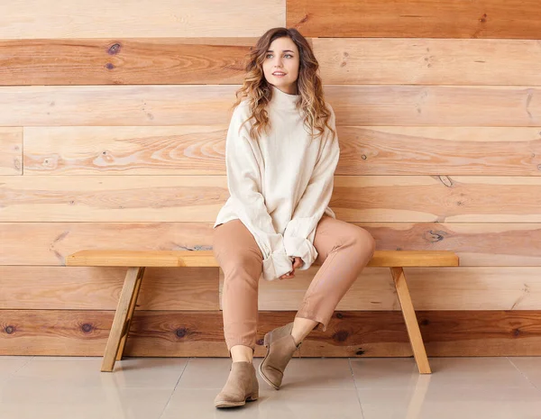 Belle jeune femme en pull chaud assise sur un banc près d'un mur en bois — Photo