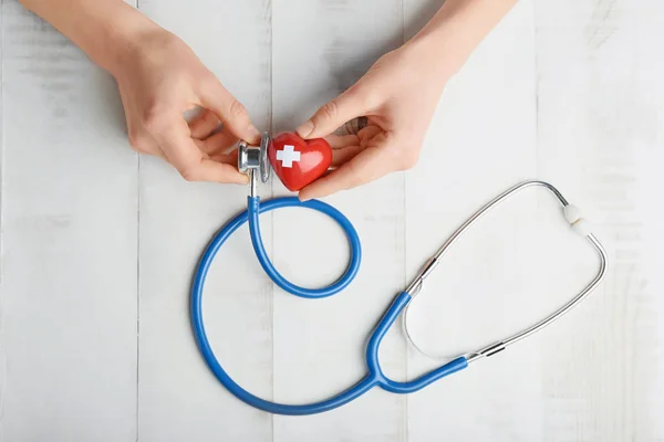 Mâini de sex feminin cu inimă roșie și stetoscop pe fundal alb din lemn. Conceptul de cardiologie — Fotografie, imagine de stoc