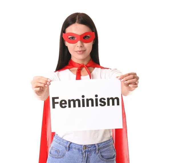 Young woman in superhero costume holding paper with text FEMINISM on white background — ストック写真