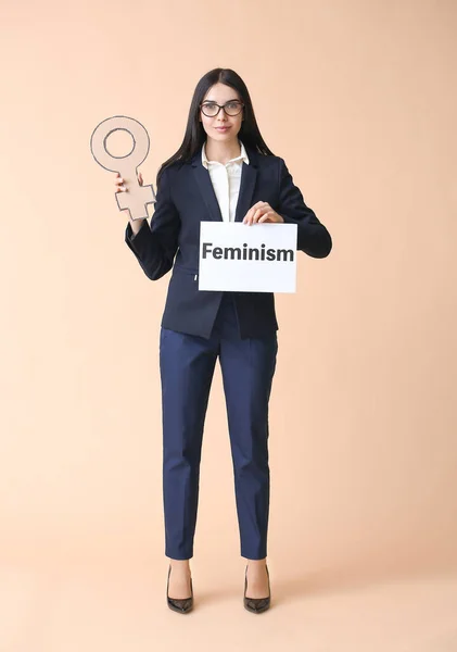 Giovane donna d'affari in possesso di simbolo di donna e carta con testo FEMINISMO su sfondo a colori — Foto Stock