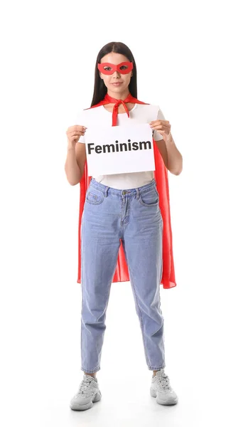 Young woman in superhero costume holding paper with text FEMINISM on white background — ストック写真