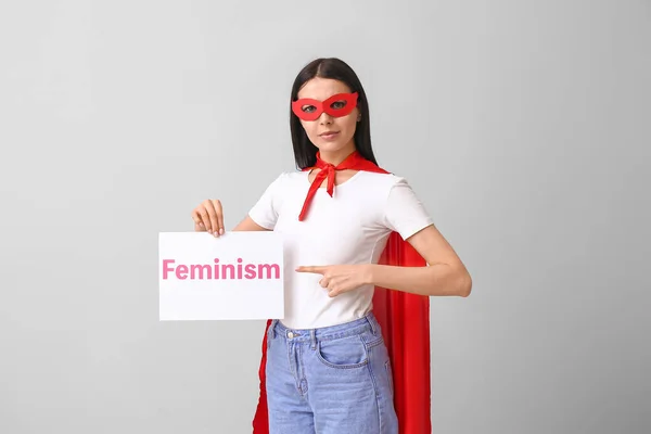 Mujer joven en traje de superhéroe sosteniendo papel con texto FEMINISMO sobre fondo gris — Foto de Stock