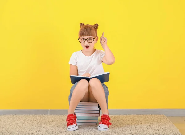 Nettes kleines Mädchen mit Büchern und erhobenem Zeigefinger sitzt in der Nähe der Farbwand — Stockfoto