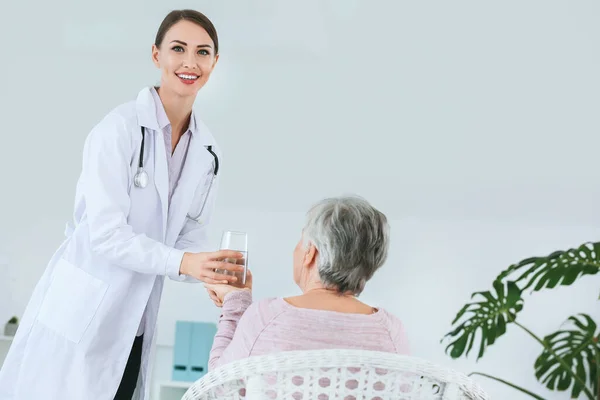 Kobieta lekarz daje starszej kobiecie cierpiącej na zespół Parkinsona szklankę wody w klinice — Zdjęcie stockowe