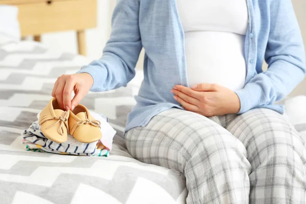 Pregnant woman with baby clothes and shoes in bedroom, closeup — Stock Photo, Image