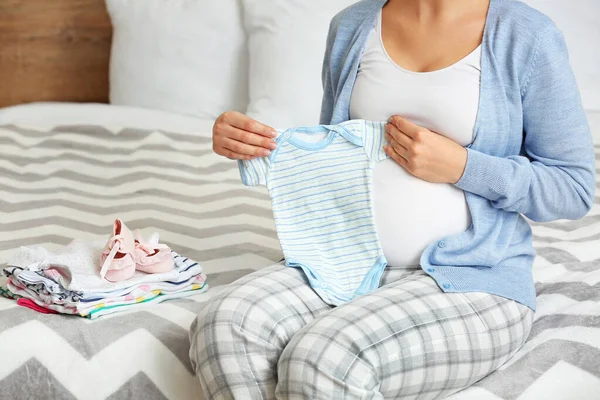 Pregnant woman with baby clothes in bedroom, closeup — Stock Photo, Image