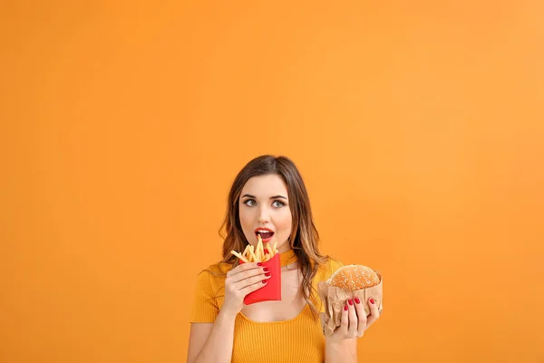 Portret van een mooie jonge vrouw met hamburger en frietjes op kleur achtergrond — Stockfoto