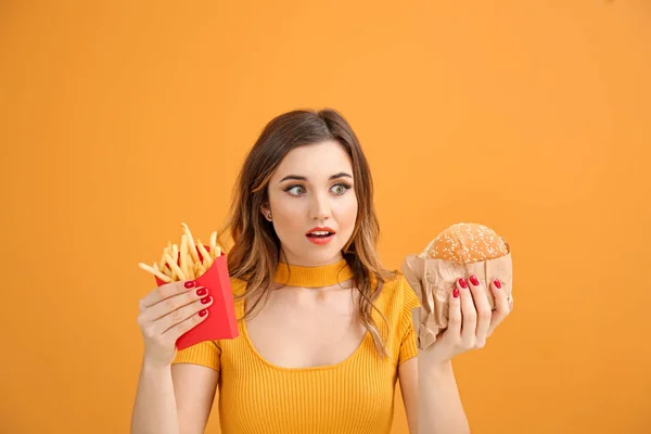 Portrait of beautiful young woman with burger and french fries on color background — Stock Photo, Image