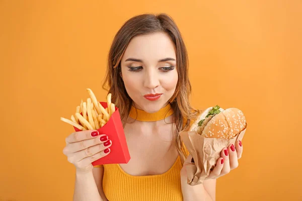 Portrait of beautiful young woman with burger and french fries on color background — Stock Photo, Image