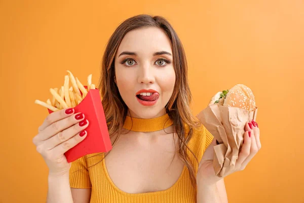 Portrait of beautiful young woman with burger and french fries on color background — 스톡 사진
