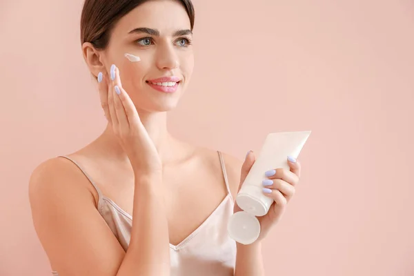 Hermosa mujer joven aplicando crema en su cara contra el fondo de color — Foto de Stock