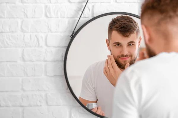 Guapo joven aplicando crema en su cara cerca del espejo en el baño — Foto de Stock