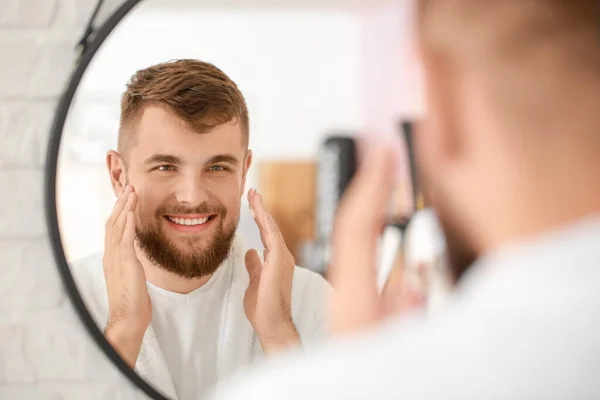 Guapo joven aplicando crema en su cara cerca del espejo en el baño — Foto de Stock