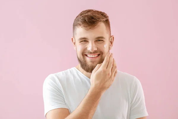 Hezký mladý muž nanášení krém na jeho tvář na barevném pozadí — Stock fotografie