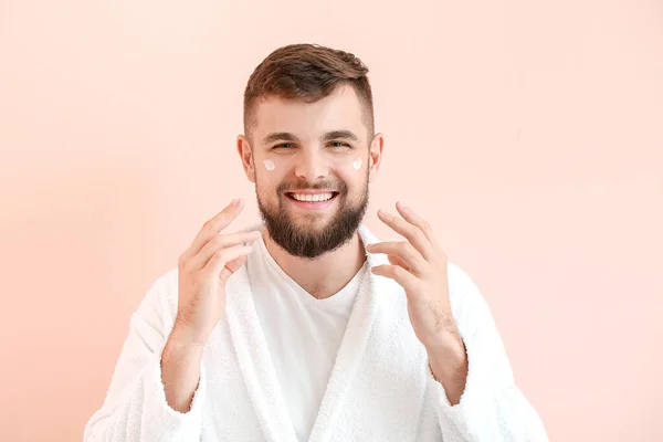 Handsome young man applying cream on his face against color background — Stock Photo, Image