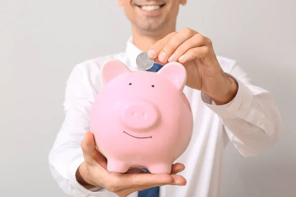 Handsome businessman putting money in piggy bank on light background — Stock Photo, Image