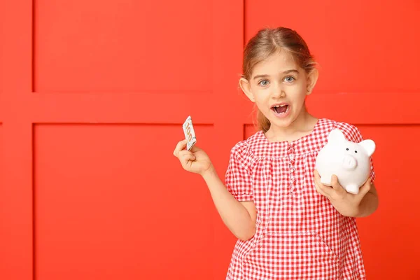 Cute girl with piggy bank and money on color background — Stock Photo, Image