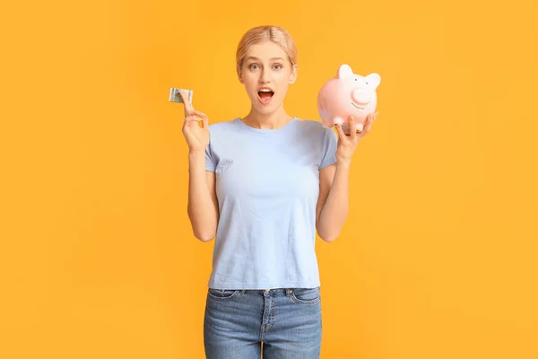 Surprised young woman with piggy bank on color background — Stock Photo, Image