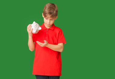 Cute boy tipping money from piggy bank on color background clipart
