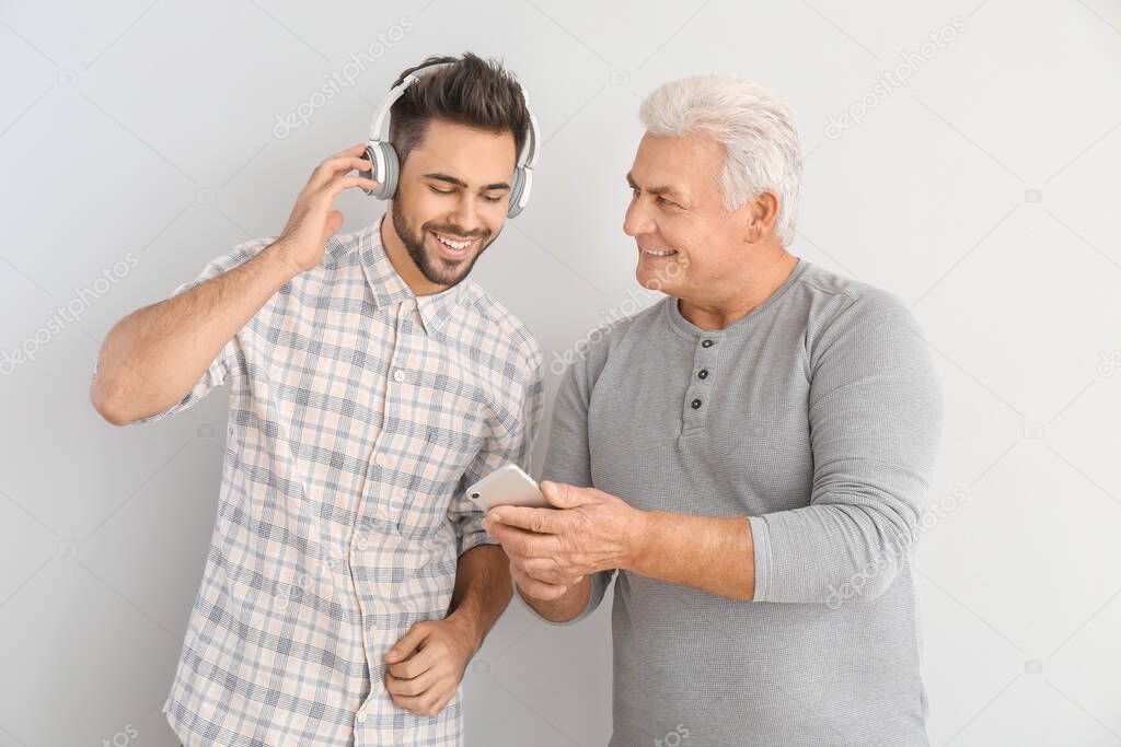 Young man and his father listening to music on light background