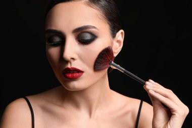 Beautiful young woman applying makeup against dark background
