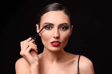 Beautiful young woman applying makeup against dark background