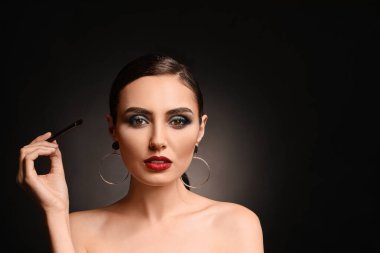 Beautiful young woman applying makeup against dark background