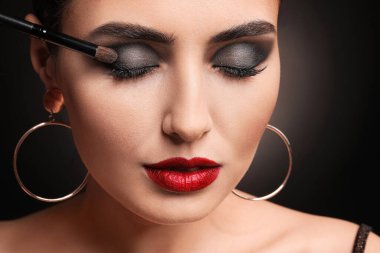 Beautiful young woman applying makeup against dark background