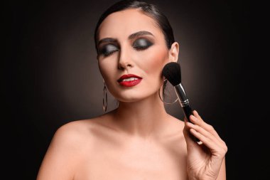 Beautiful young woman applying makeup against dark background