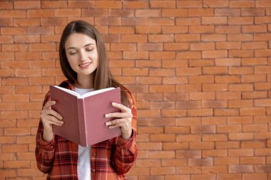 Güzel genç bir kadın tuğla arka planda kitap okuyor.