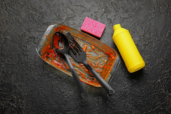 Dirty baking dish, kitchen utensils, detergent and sponge on dark background