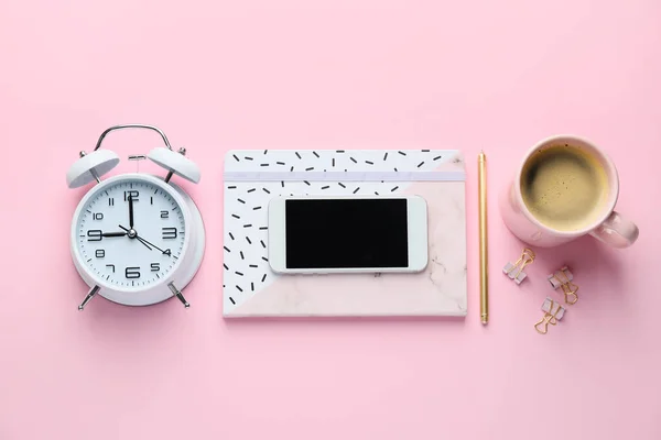 Mobile phone with stationery, alarm clock and cup of coffee on pink background