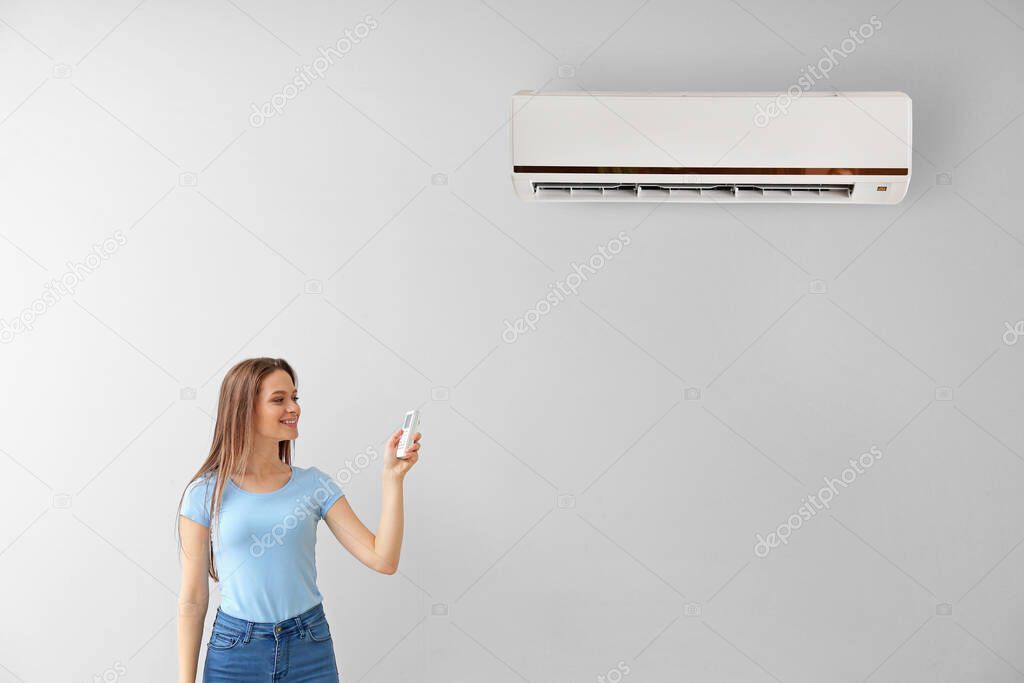 Young woman switching on air conditioner on light wall