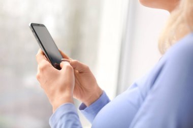 Pencerenin yanında cep telefonu olan genç bir kadın.