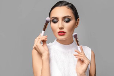 Gri arkaplanda makyaj fırçaları olan güzel bir genç kadın.