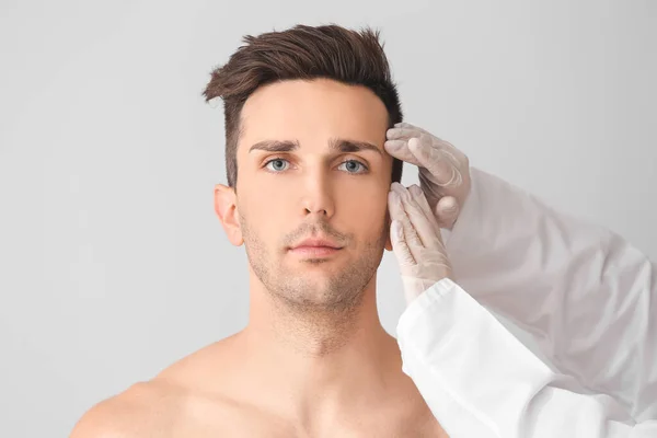 Plastic surgeon touching face of young man on light background
