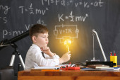 Netter kleiner Junge mit glühender Glühbirne im Physikunterricht im Klassenzimmer
